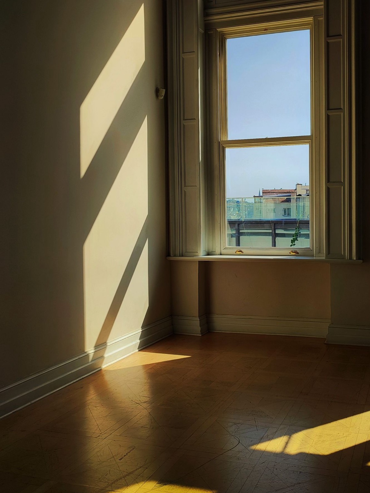 An empty room with a tall sash window, afternoon light falling in long shapes across bare floorboards.