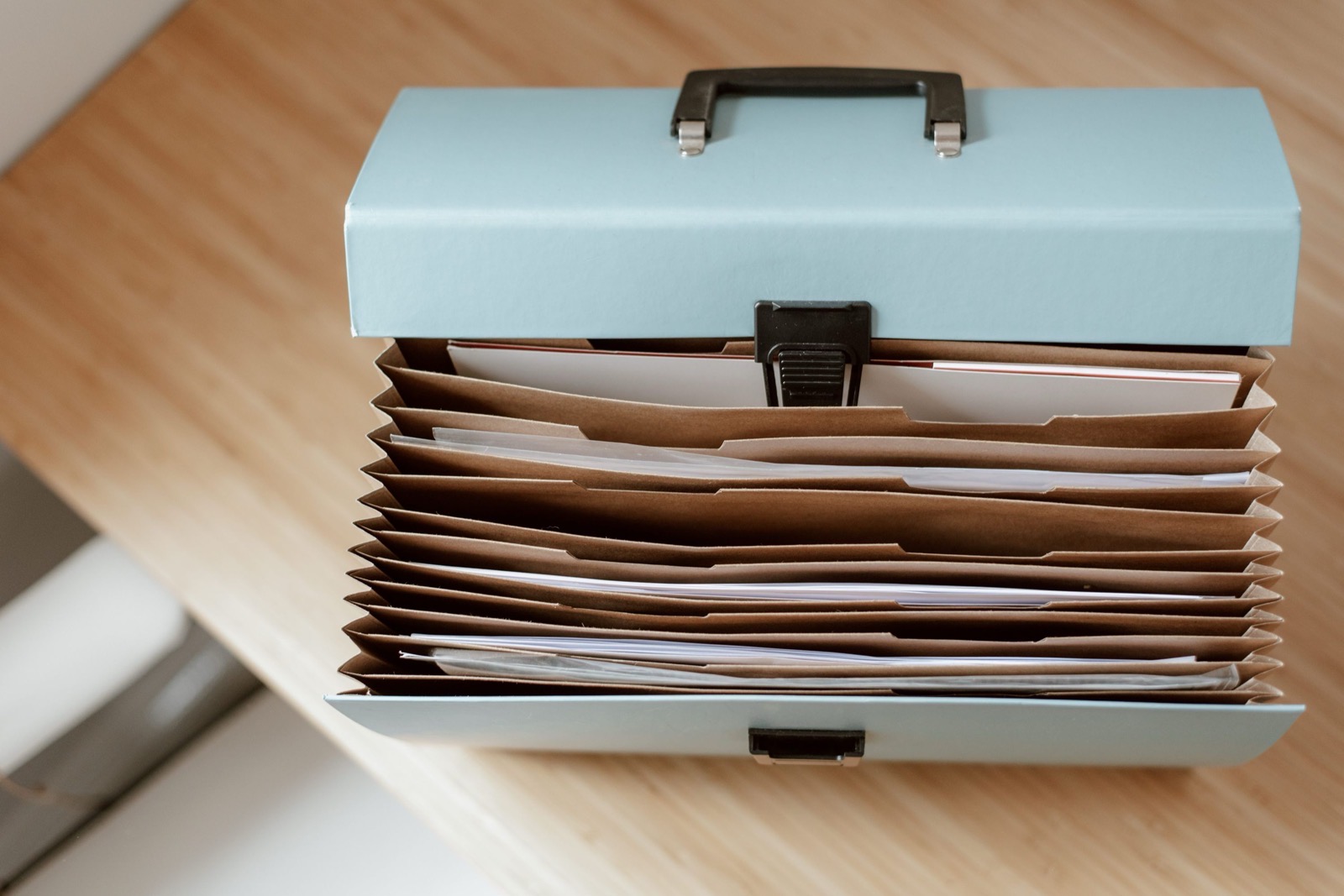 An open concertina file of kraft paper dividers packed with documents, standing on a pale timber bench in soft daylight.