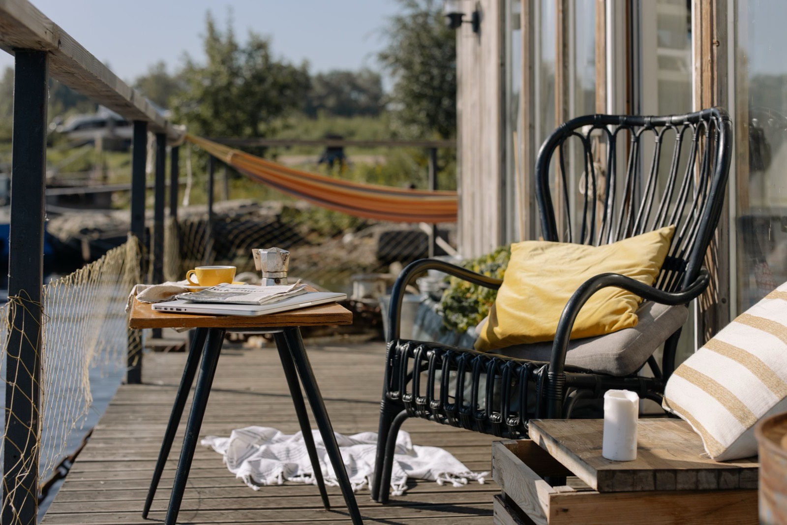 A small side table on a timber deck holding a coffee cup and a spread of papers, a cane chair beside it and open garden beyond the railing.